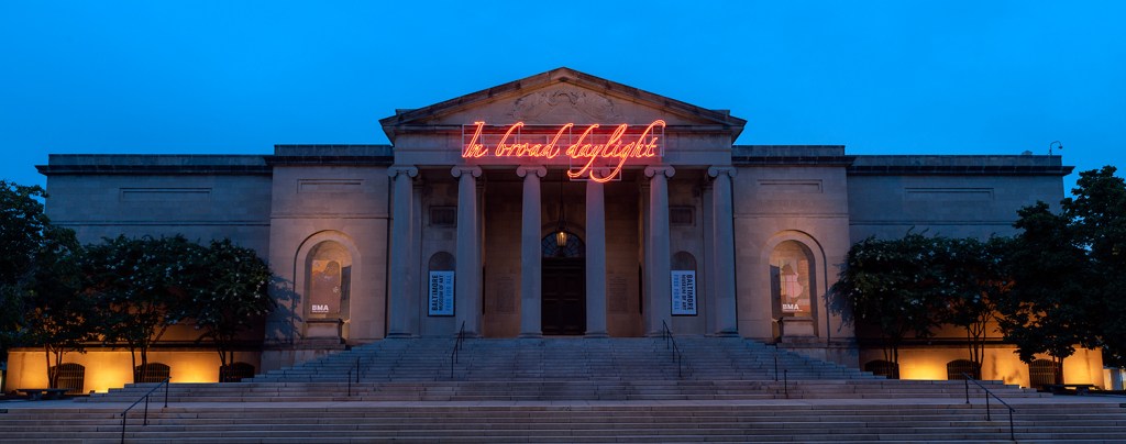 An evening photo of the six-columned entrance to the Baltimore Museum of Art, featuring an orange neon light sculpture by Travares Strachan proclaiming, "In broad daylight"