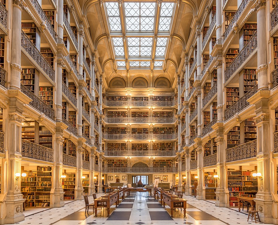 A photo featuring the inside of the George Peabody Library at Johns Hopkins University, with patterned marble floors, long wooden desks to study, a sky-lit roof, and views of five open floors of library stacks and ornate architecture