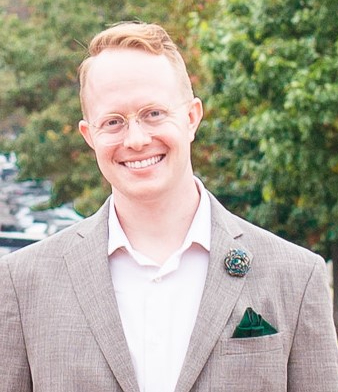 A square photo of Dr. Jordy in front of a forest background wearing a beige sport coat with dark green accents. He has strawberry blonde mid-length hair and is wearing vintage glasses. 